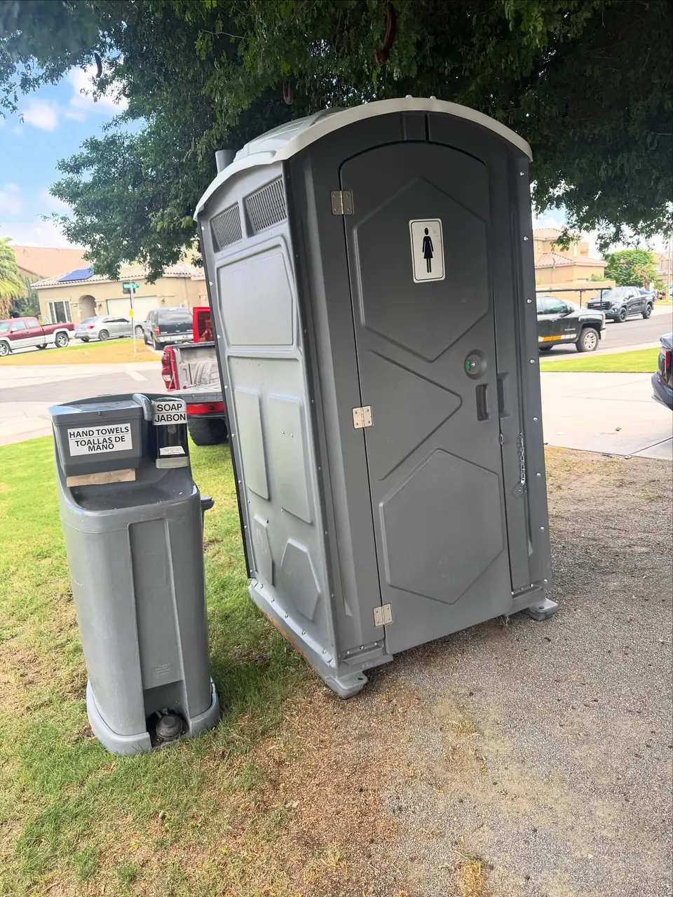 Gray portable toilet with a women's sign next to a hand-wash station on a residential lawn