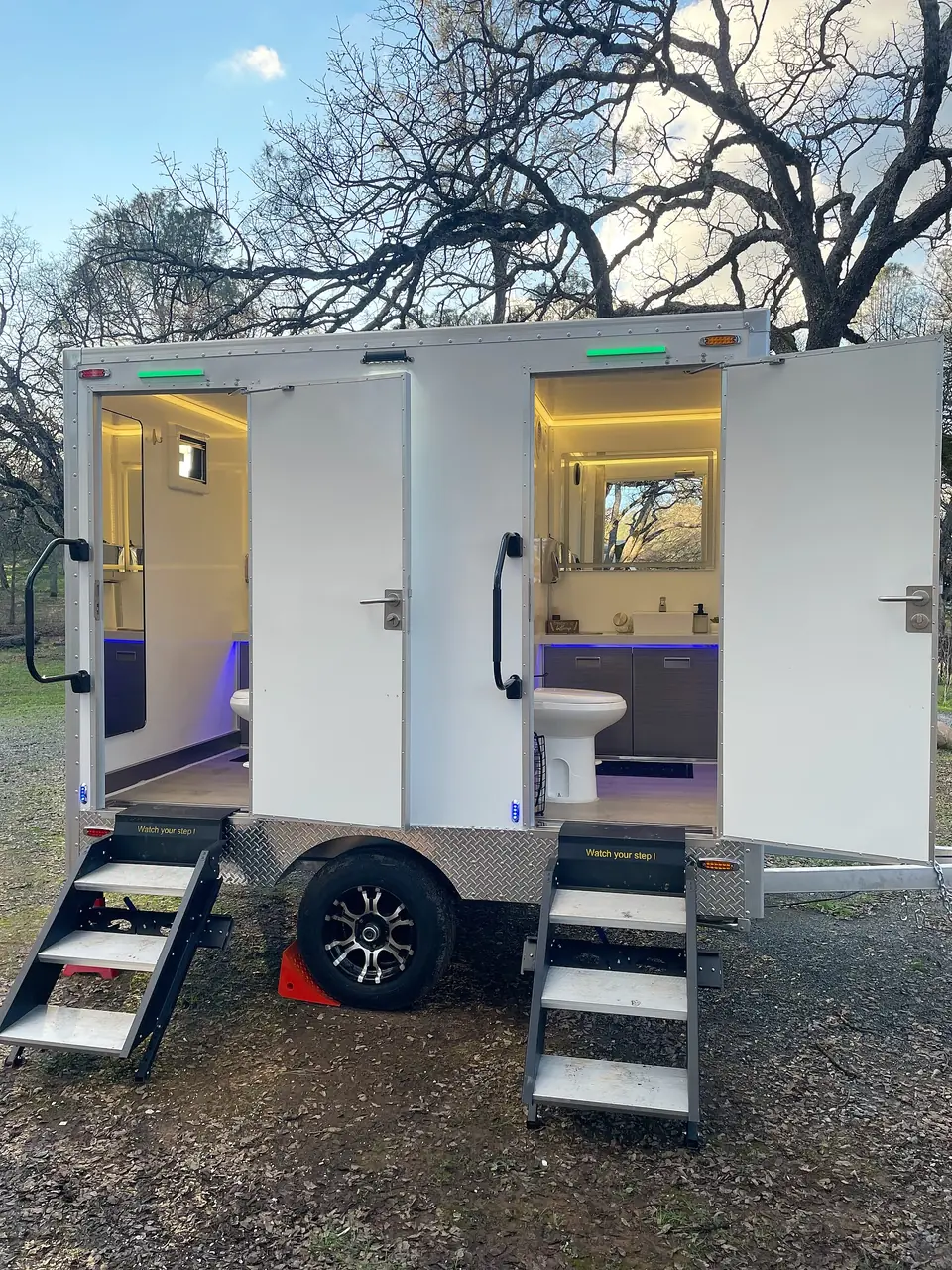 Two-stall restroom trailer with lights on and doors open at an outdoor event