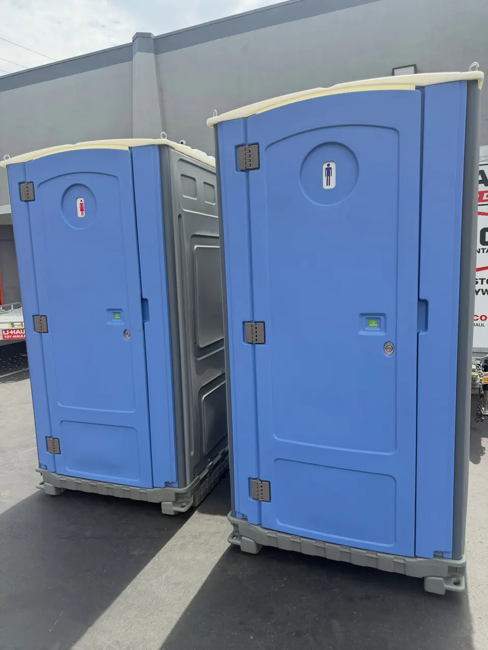 Two blue porta potties side by side on asphalt next to a commercial building